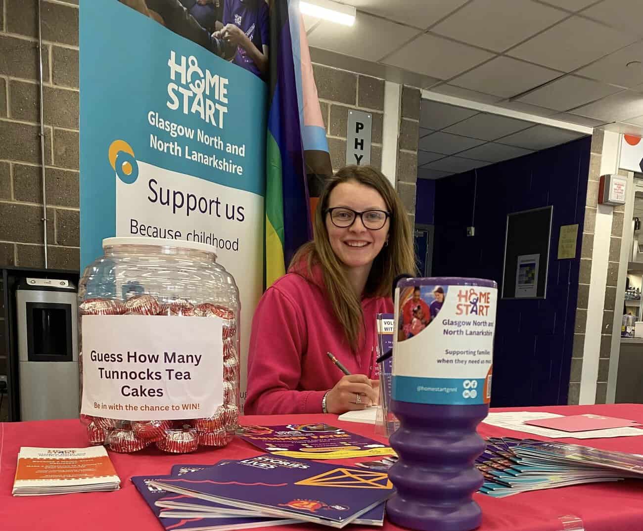 Image of Rachel sitting on a table with Home-Start promotional material, and and pop up banner behind her.