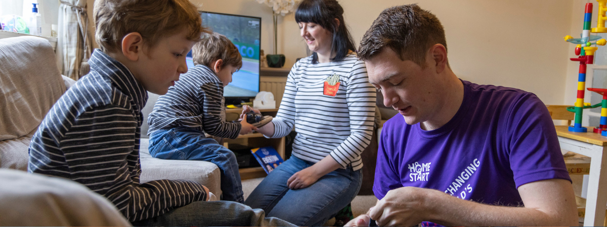 Image of volunteer helping a young boy put his shoes on, mum is in the background helping the other young boy with his shoes