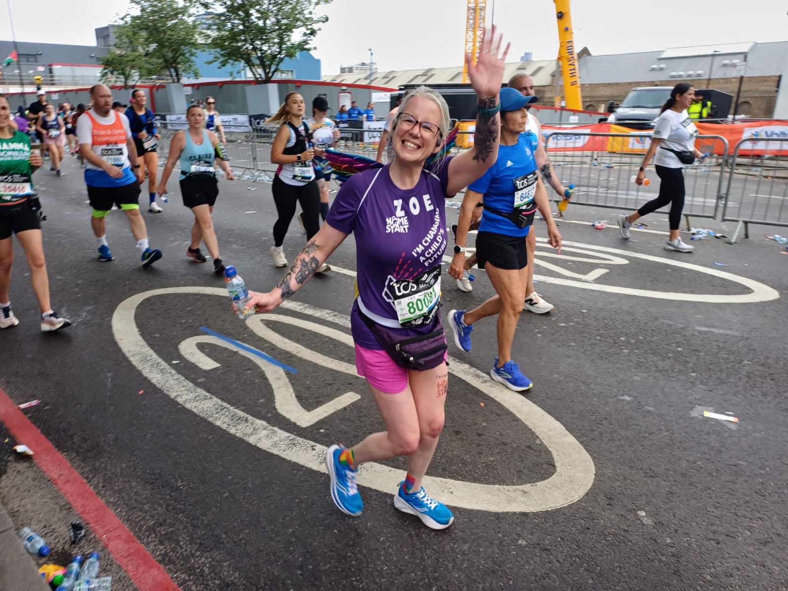 Image of a women running the London Marathon, smiling and waving. She is wearing a purple Home-Start T-Shirt
