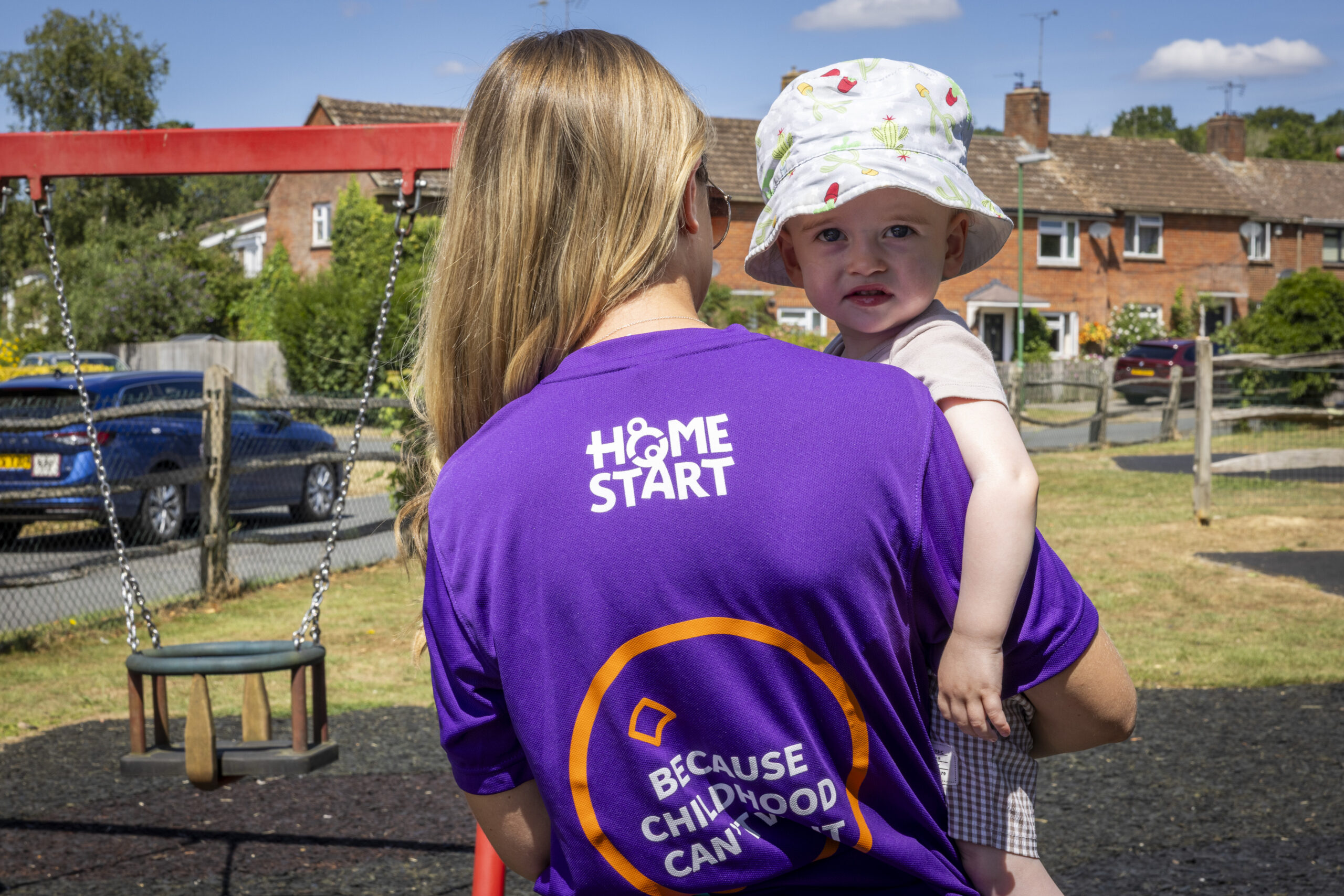 Image of a Home-Start Volunteer with their back to camera wearing a purple t-shirt with Home-Start logo. Holding baby who is wearing a sun hat and looking at the camera.