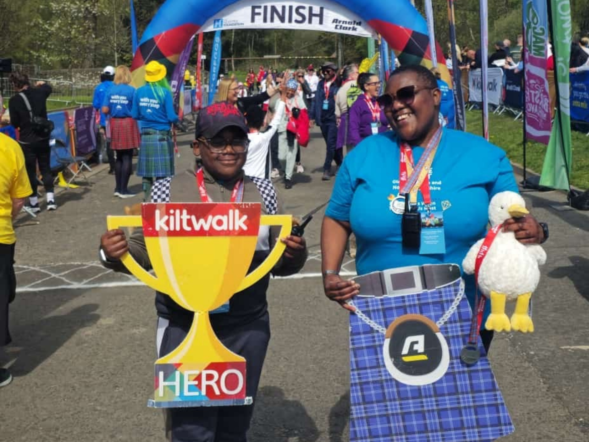 Image of Leah our volunteer and her son at the end of the Kiltwalk holding a Kiltwalk cut-out trophy and a kilt.