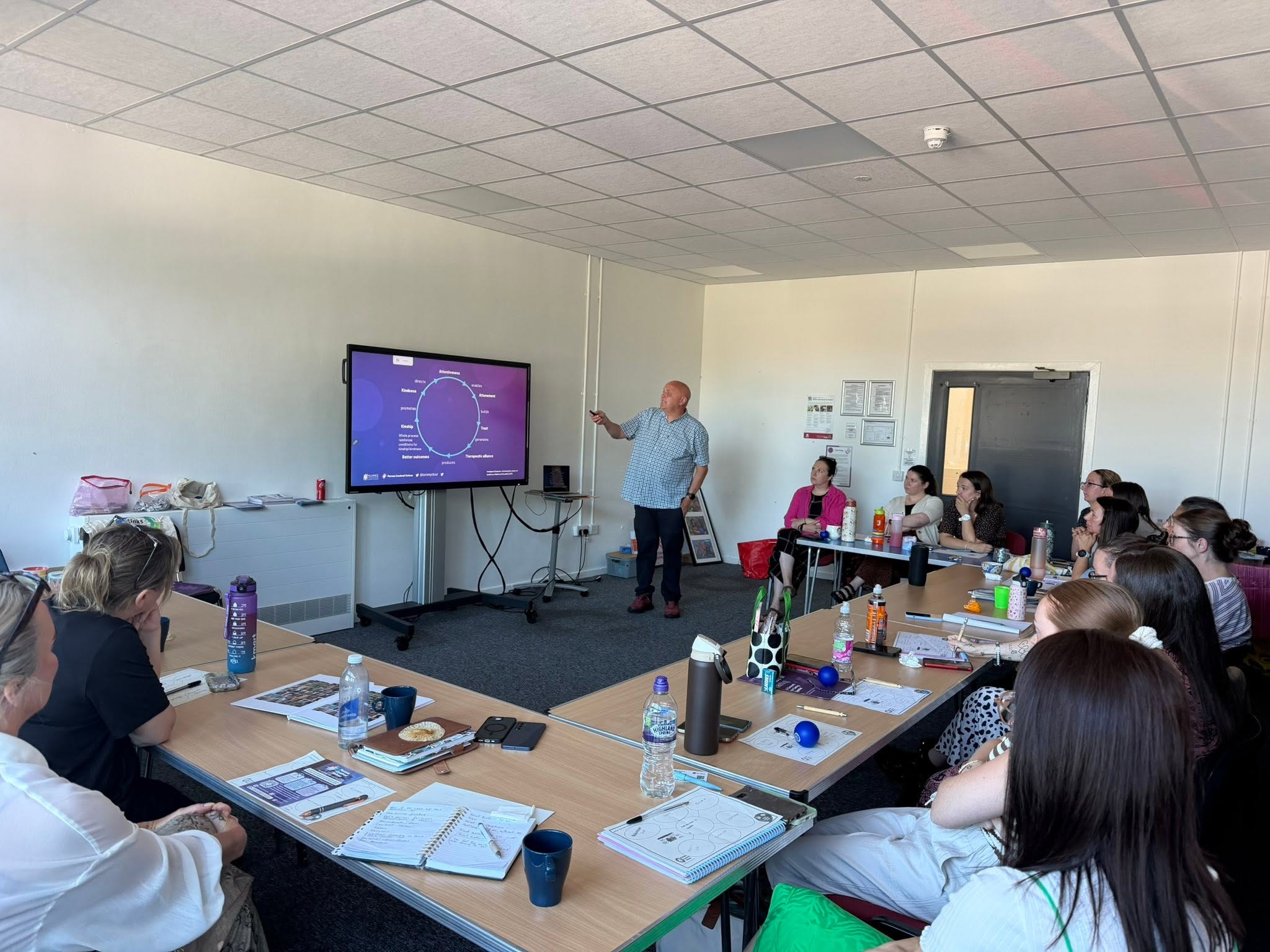 Image of the team in a meeting room sat around tables watching Tommy Whitelaw tood at the front delivering his person centre talk on kindness, he is pointing at a large monitor.