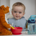 Image of baby sitting in a high chair, with a bowl and spoon, a tiger teddy and a water bottle.